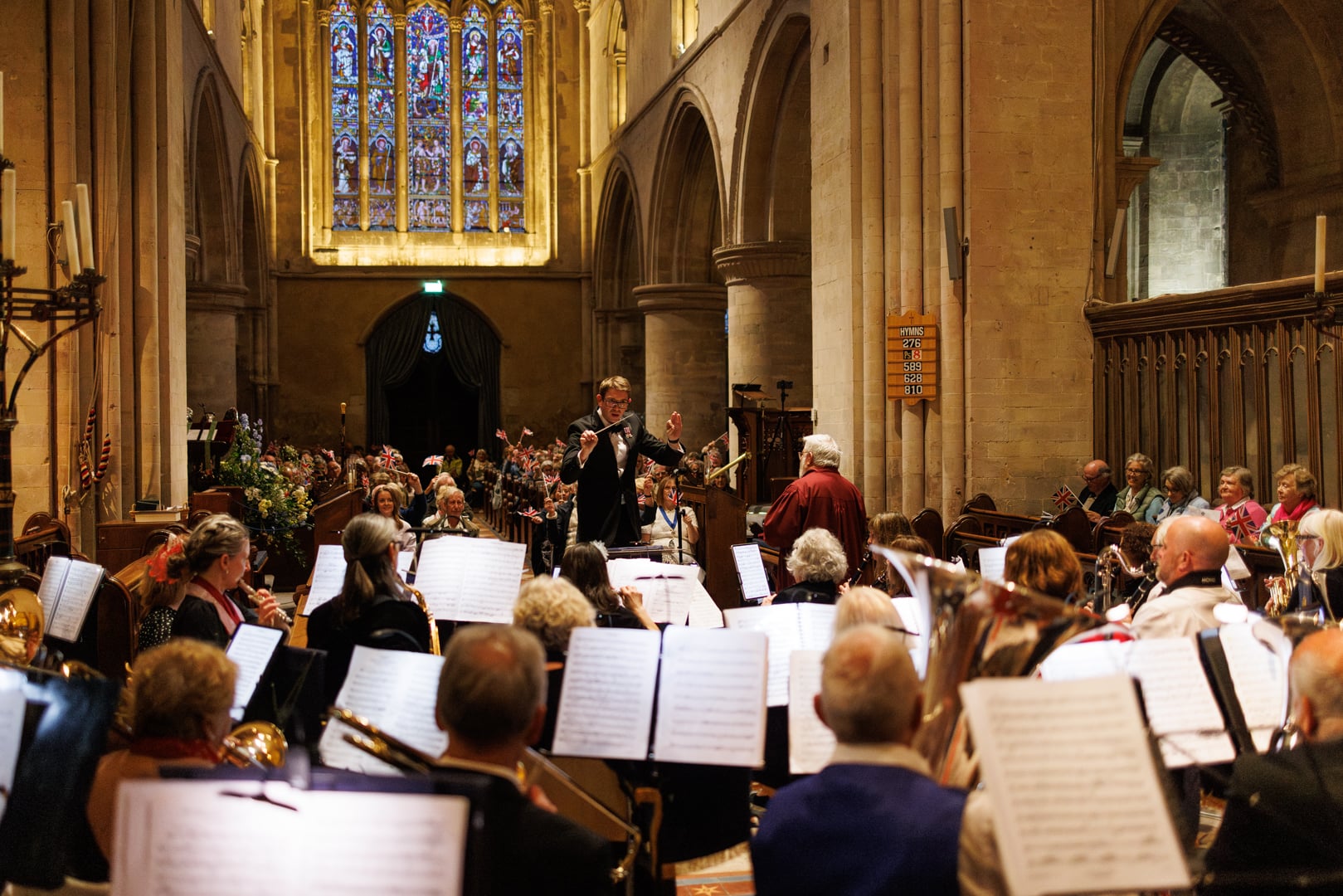 James Farthing conducting an orchestra in a cathedral nave, with the audience waving flags beneath a stained-glass window
