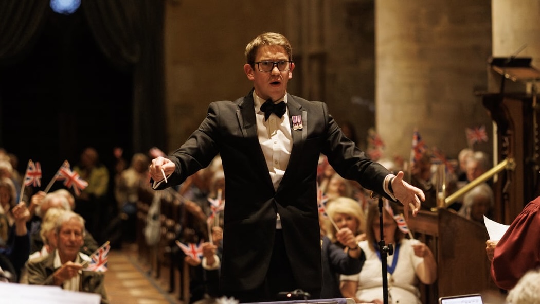 James Farthing conducting in a cathedral, arms outstretched, with an audience waving Union Jack flags behind him